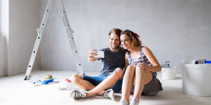 Två glada personer tar en selfie sittandes på  ett golv i ett rum som renoveras.